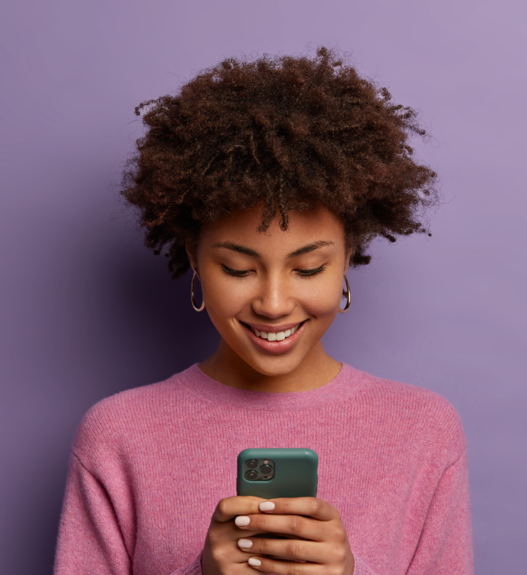 Smiling young woman in a pink sweater looking at smartphone against a purple background — symbolizing digital engagement and social media marketing.